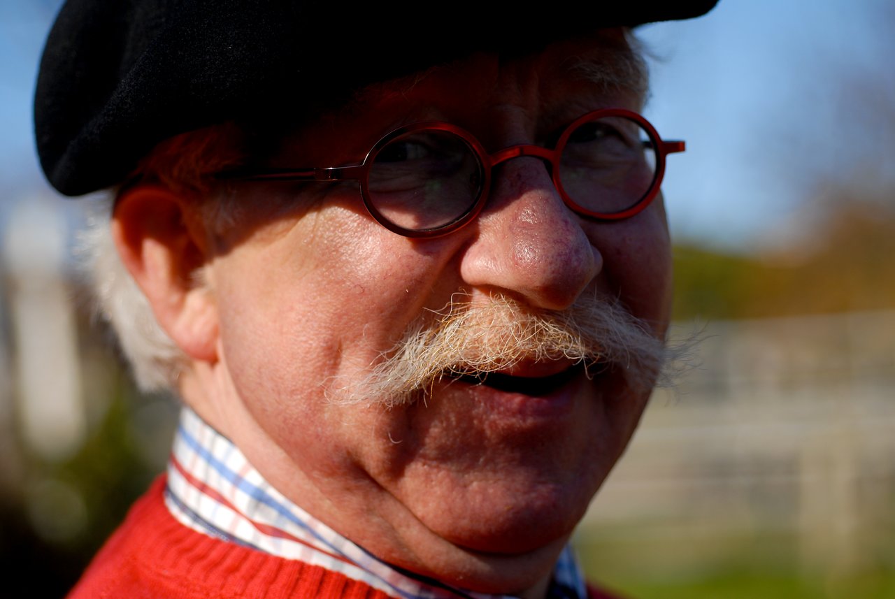 An older man with a mustache, wearing round glasses, a black beret, and a red sweater, smiling outdoors.