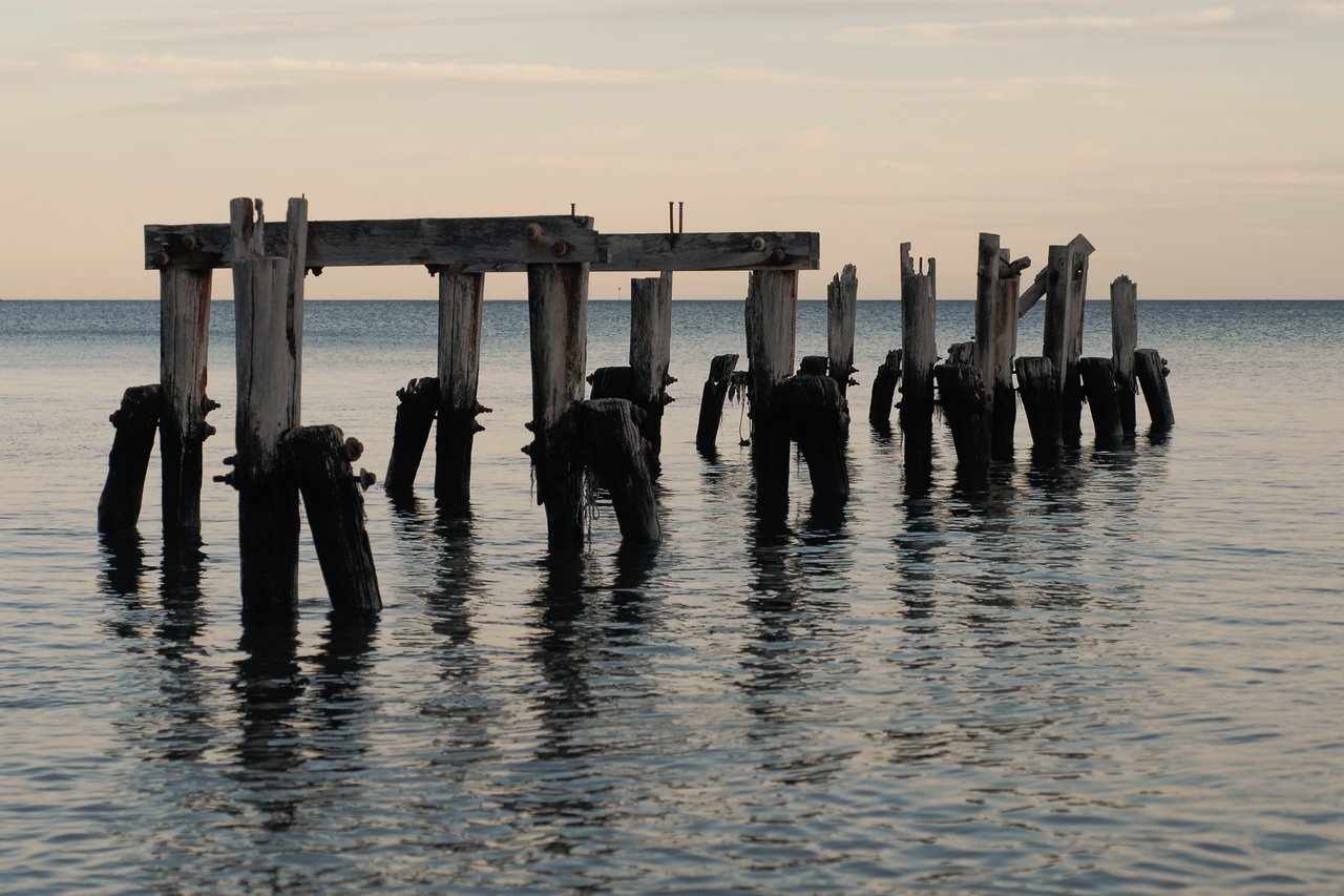 Partially collapsed wooden pier with broken posts standing in calm water, extending toward the horizon under a soft sky.