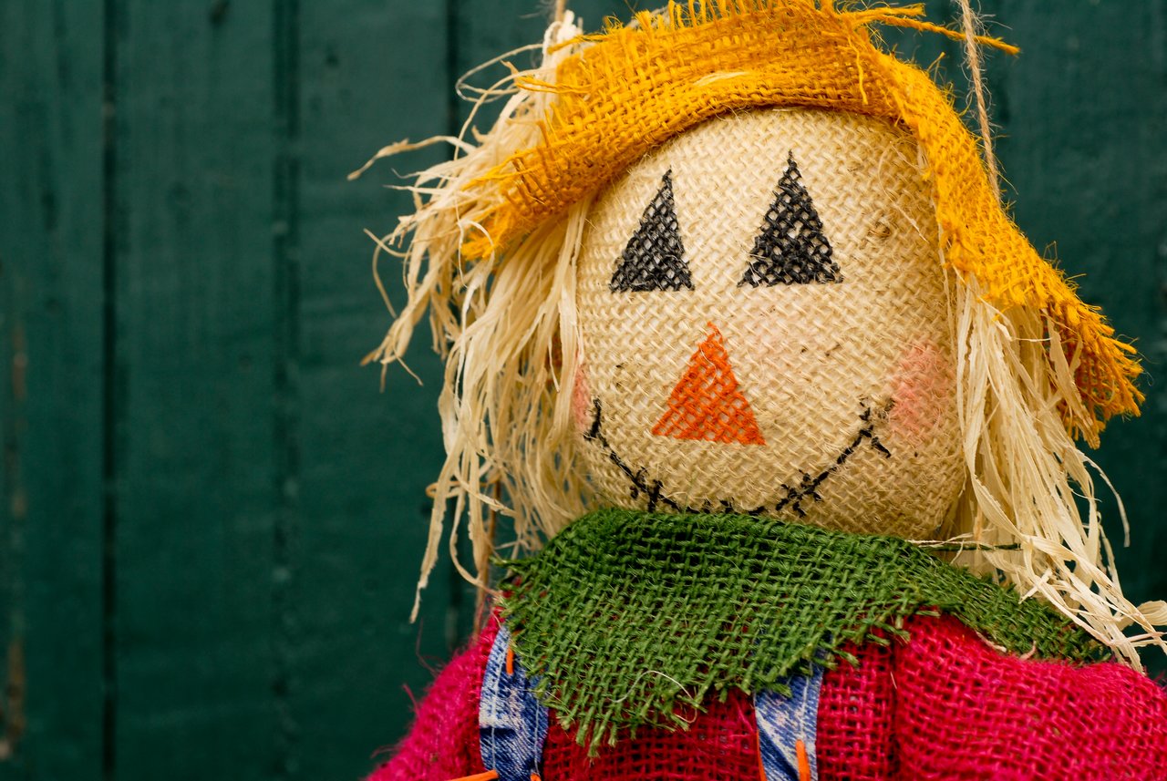 A burlap scarecrow puppet with a stitched face, straw hair, and a yellow hat, wearing colorful fabric clothing.