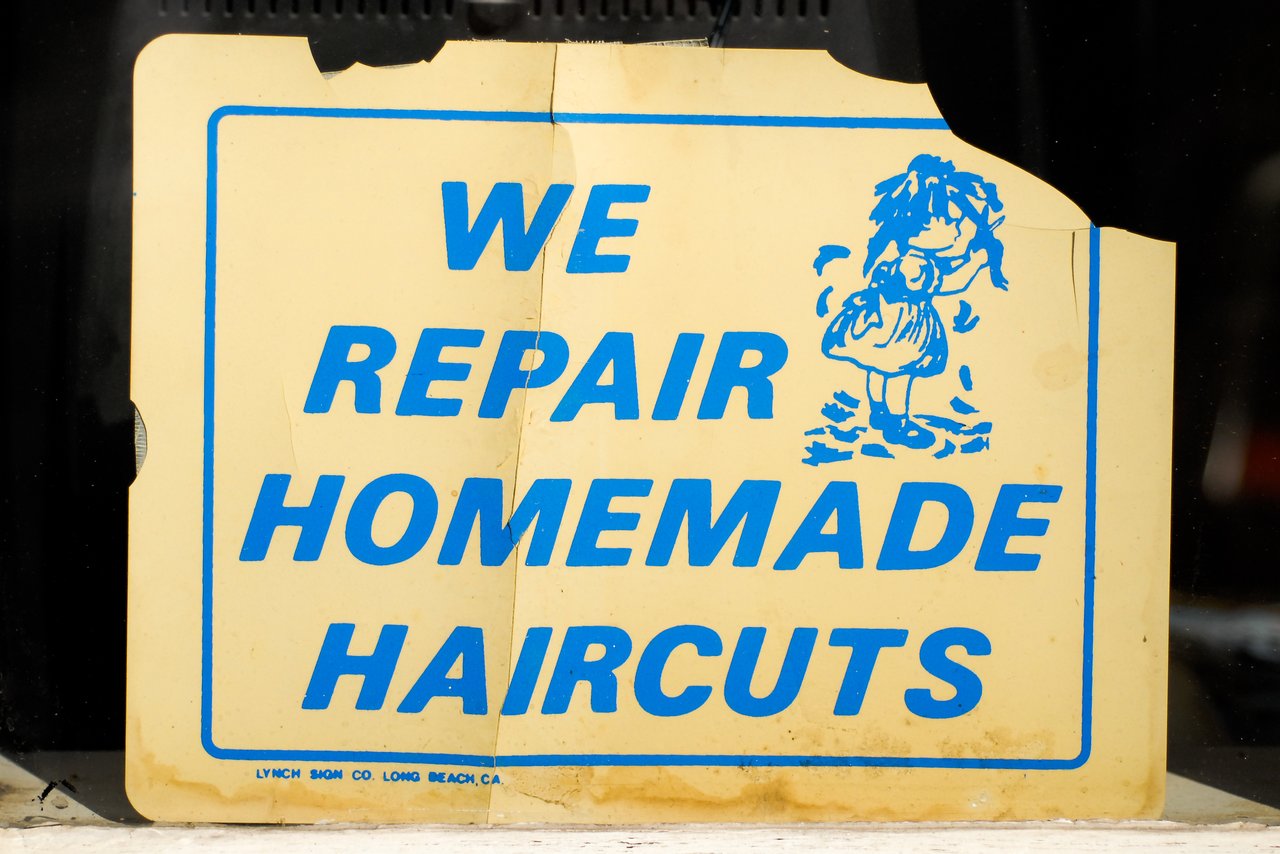A worn sign in a window reads, "We Repair Homemade Haircuts", with an illustration of a child with messy hair.