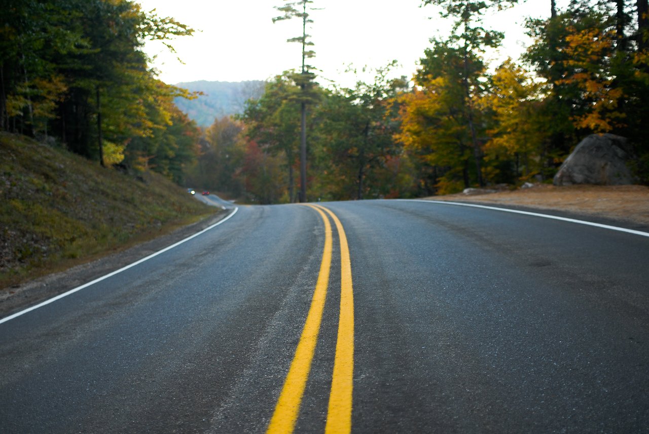 A two-lane road with yellow dividing lines curves through a wooded area with trees showing autumn colors.