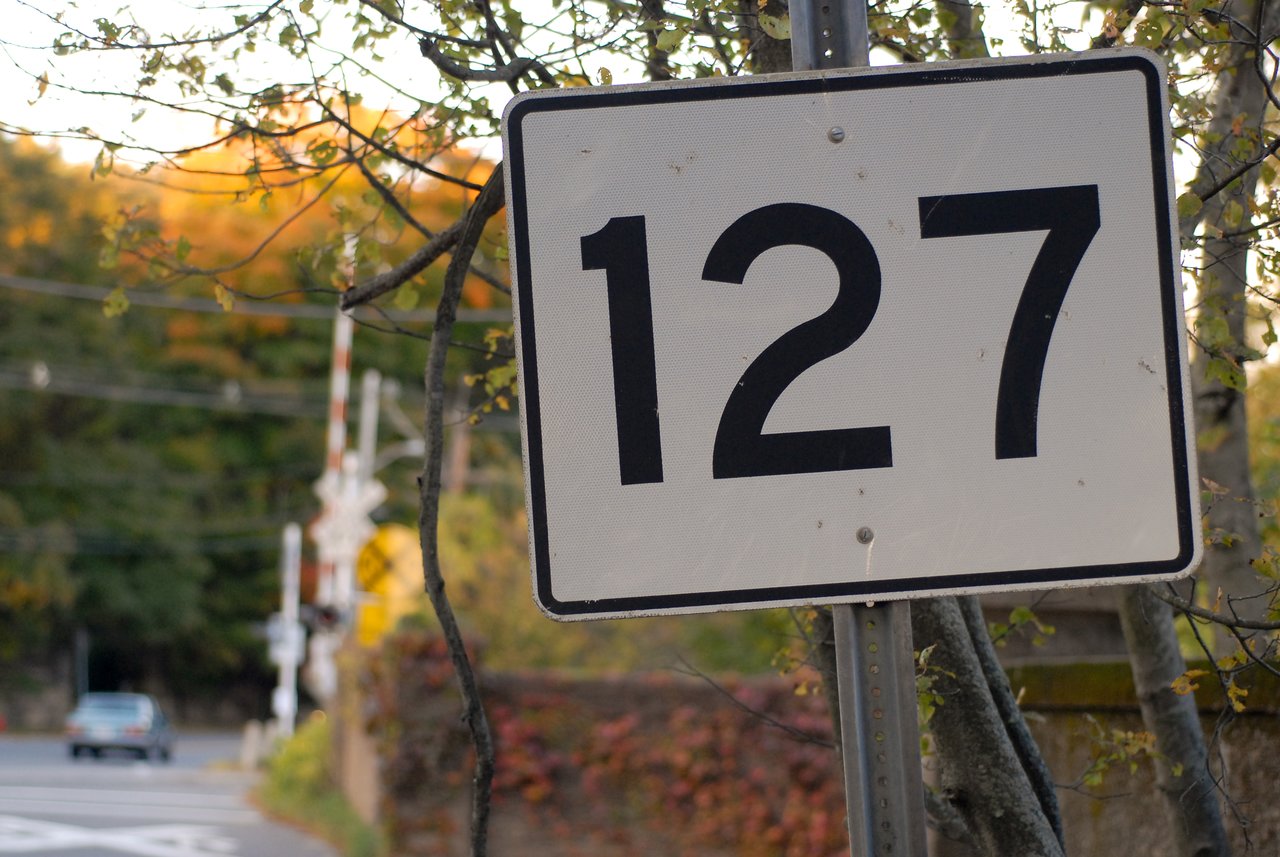 A road sign with the number 127 stands near trees, with a car and railroad crossing in the background.
