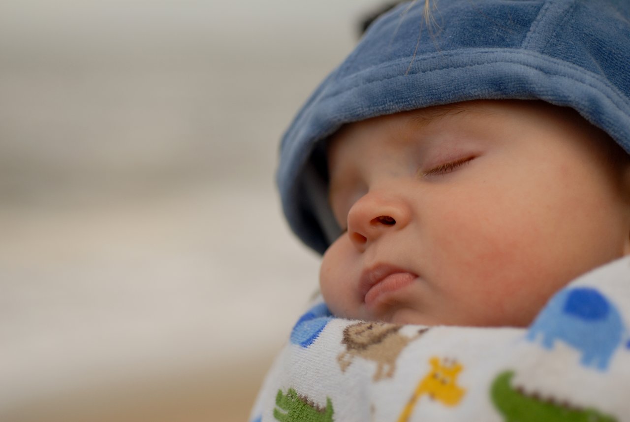 A baby sleeps peacefully, wrapped in a patterned blanket and wearing a soft blue hat.
