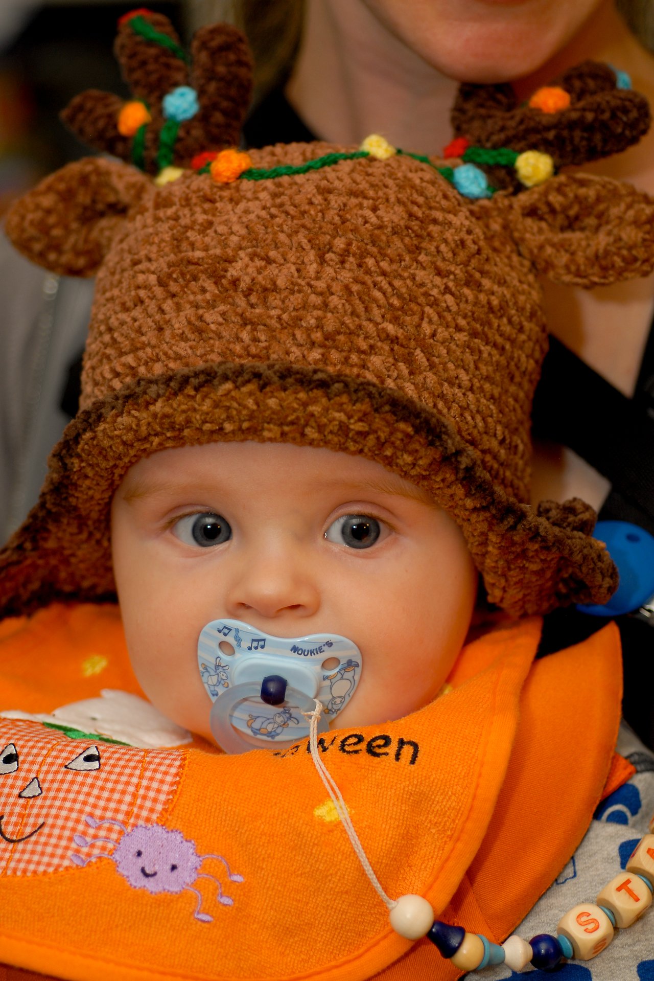 A baby wearing a reindeer-themed hat and an orange Halloween bib sucks on a pacifier while being held.