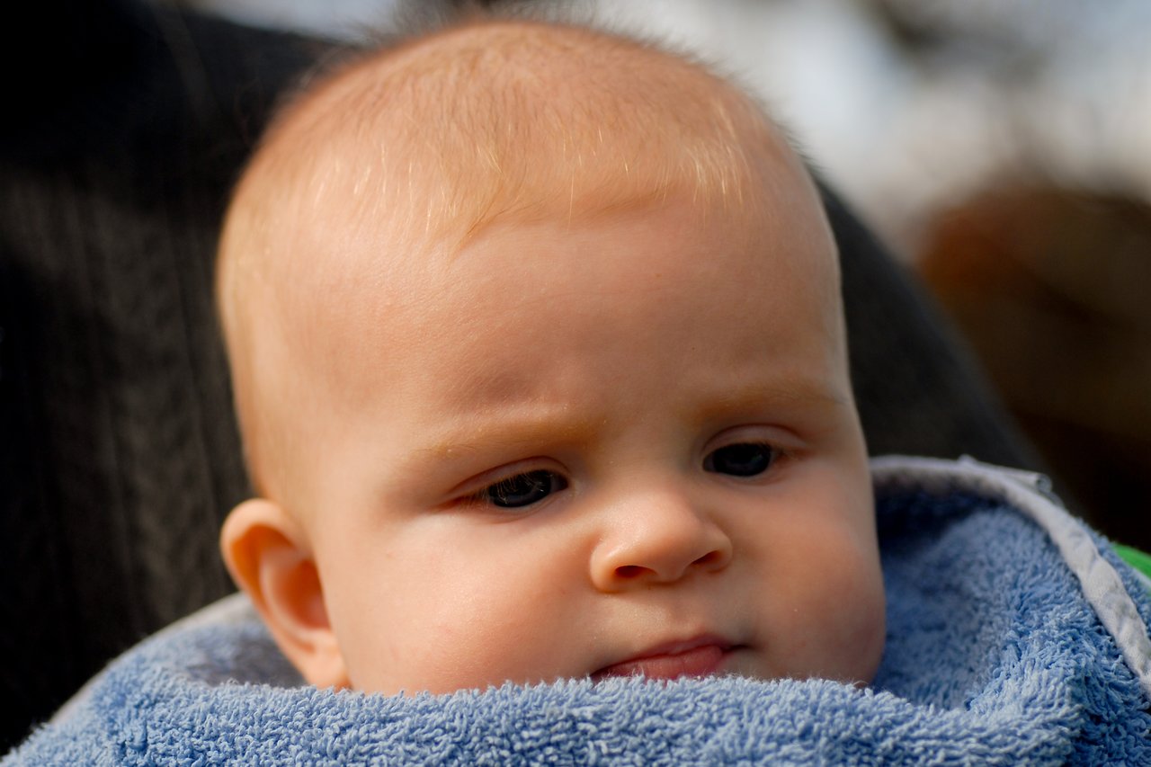A baby with light skin and short hair is wrapped in a blue towel, looking downward with a serious expression.