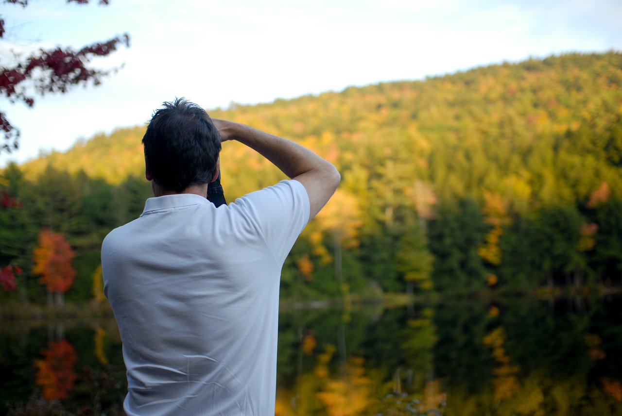 A man in a white shirt takes a photo of a scenic autumn landscape with a camera.