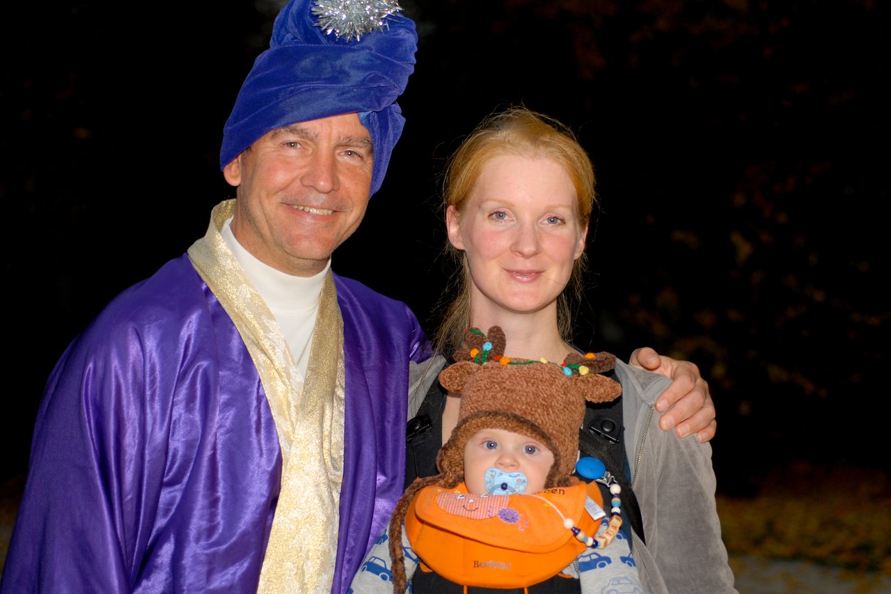 A man in a costume, a woman, and a baby in a reindeer hat pose together and smile.