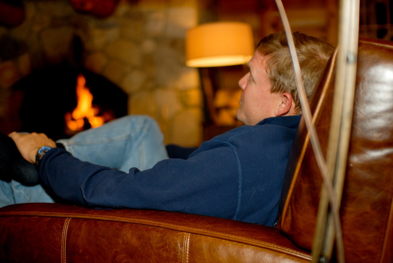 A man in a blue sweater reclines in a leather chair with his eyes closed near a fireplace.