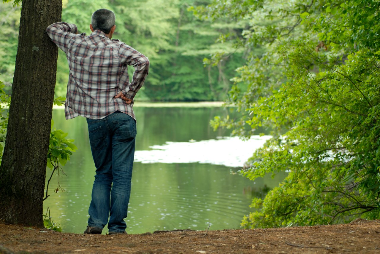 A man in a plaid shirt leans against a tree, looking at the lake with a thoughtful posture.