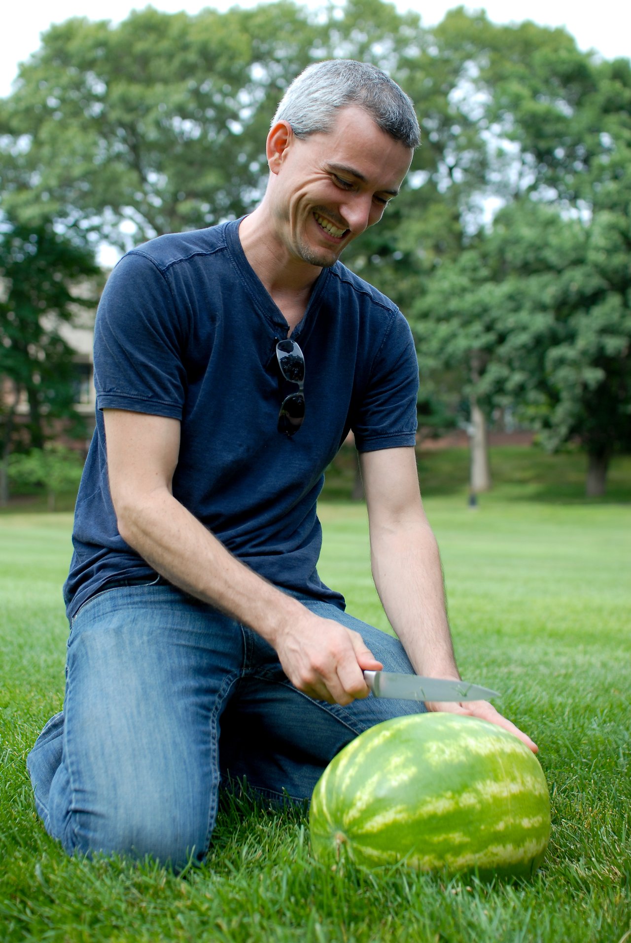 A man kneels on grass, smiling while cutting a watermelon with a knife.