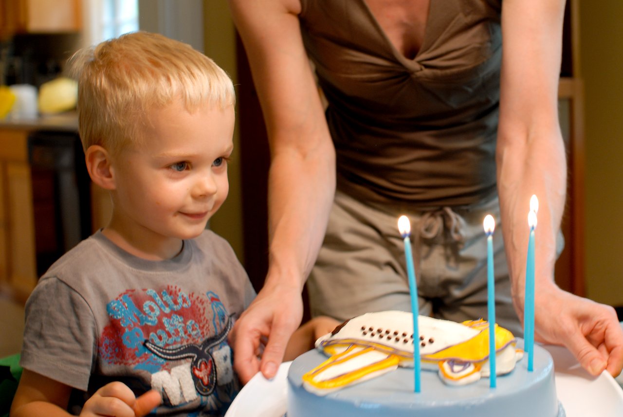 A child smiles as an adult presents a blue birthday cake with an airplane design and lit candles.