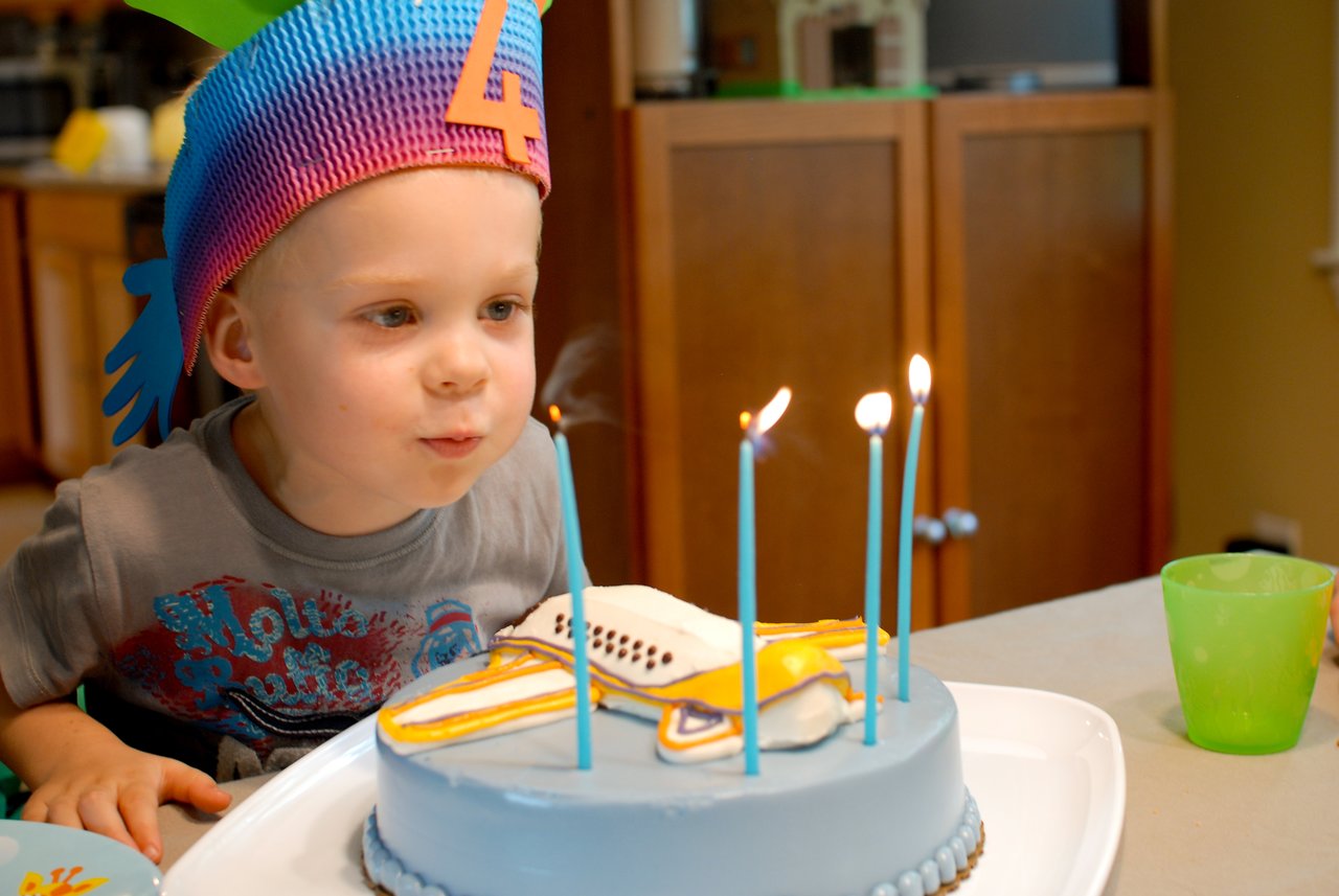 A young child wearing a colorful hat blows out candles on a blue birthday cake with an airplane design.