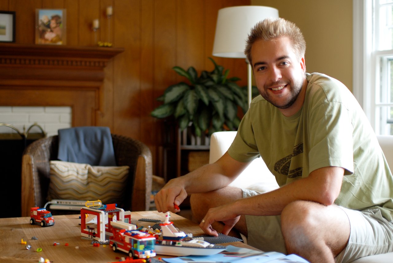 A man sitting on a couch smiles while assembling a LEGO fire station set on a wooden table.