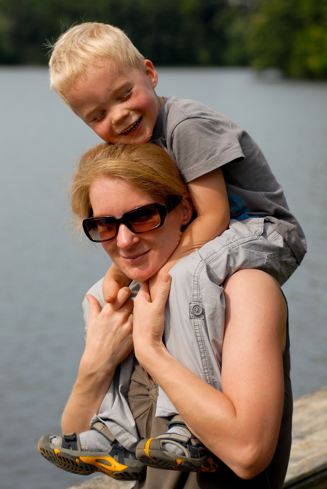 A woman carries a smiling young boy on her shoulders near a body of water.