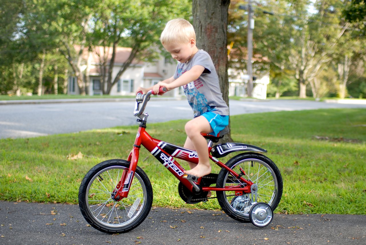 A young child rides a new red bike with training wheels on a paved path.
