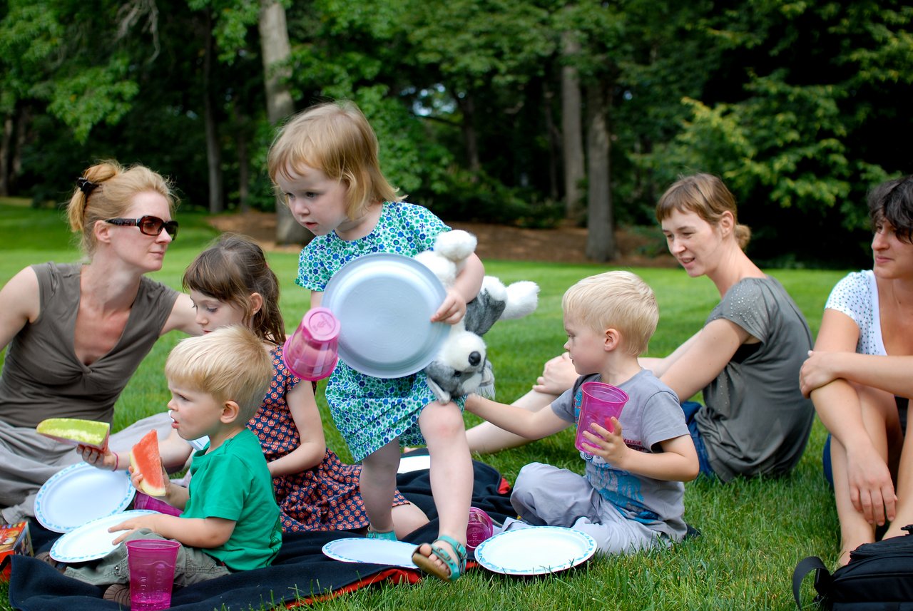 Children and adults sit on a picnic blanket, eating watermelon and holding plates and cups in a grassy park.