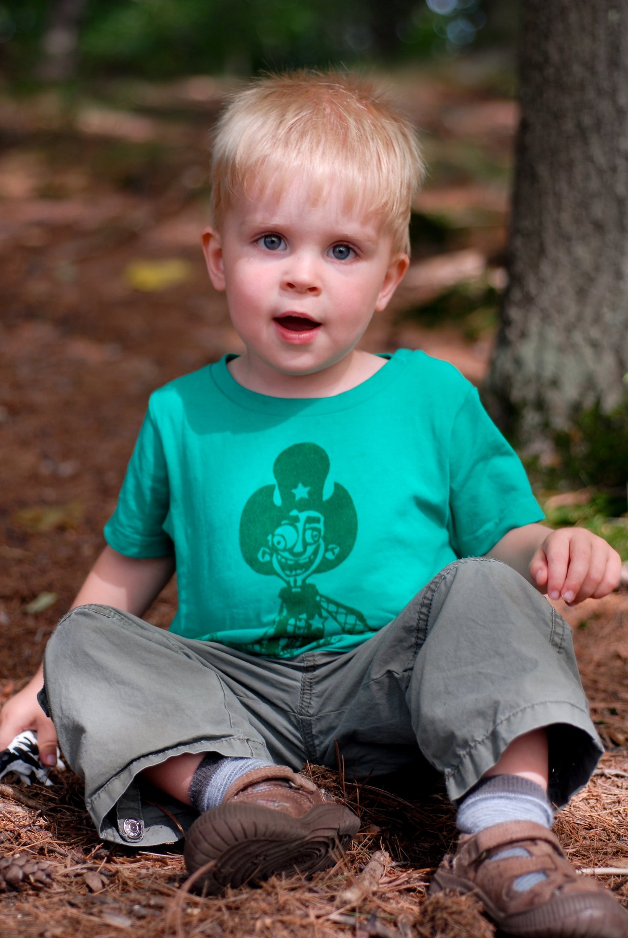A young child with blonde hair sits on the ground outdoors, wearing a green shirt and looking at the camera.