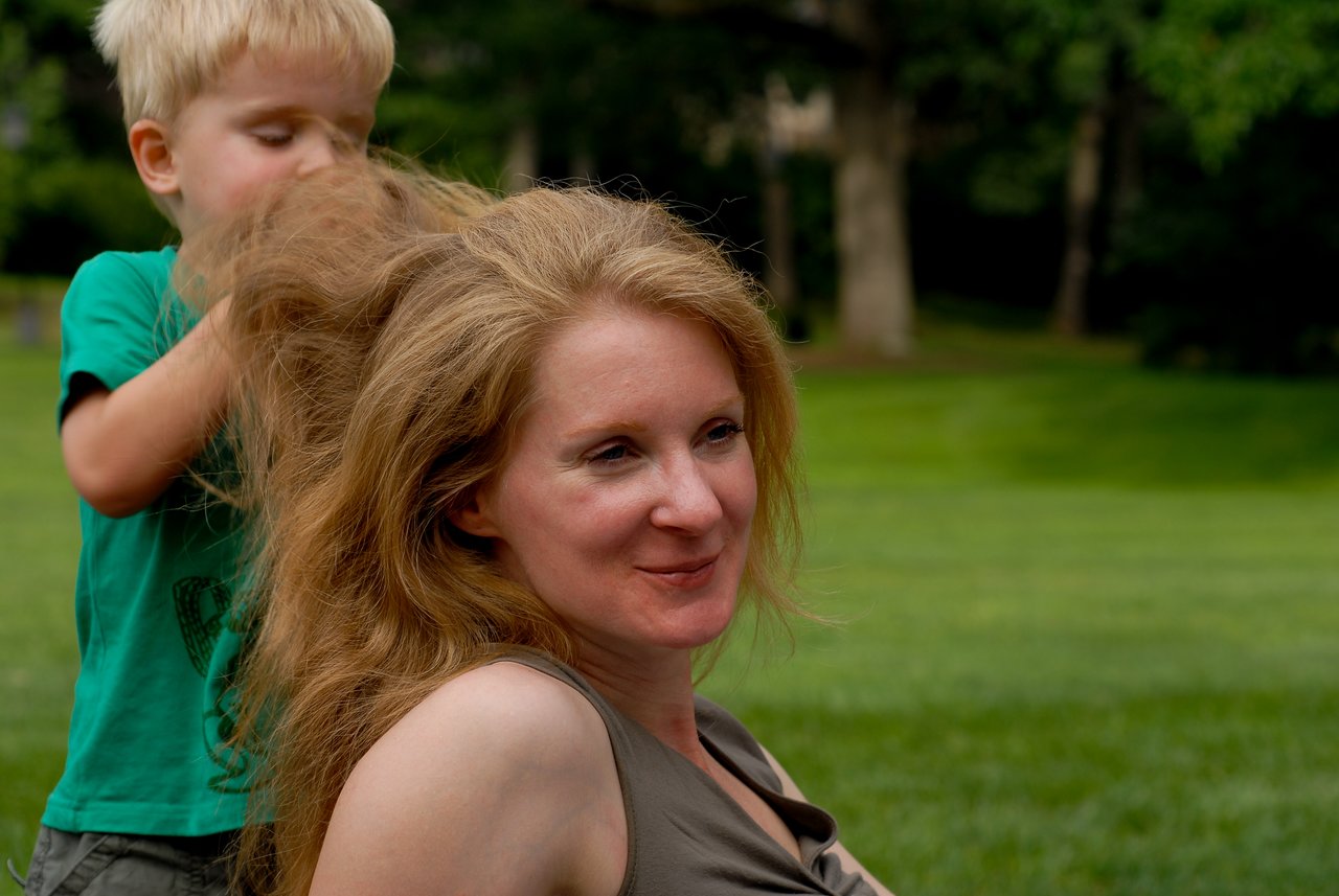 A young boy playfully styles a smiling woman's hair outdoors in a grassy park.