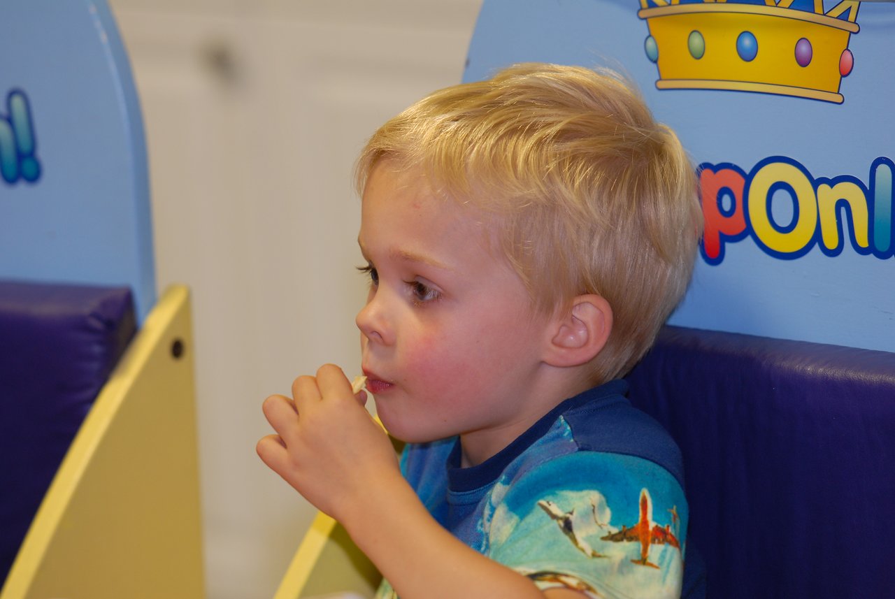 A young child with blonde hair sits in a booth, eating pizza and looking to the side.