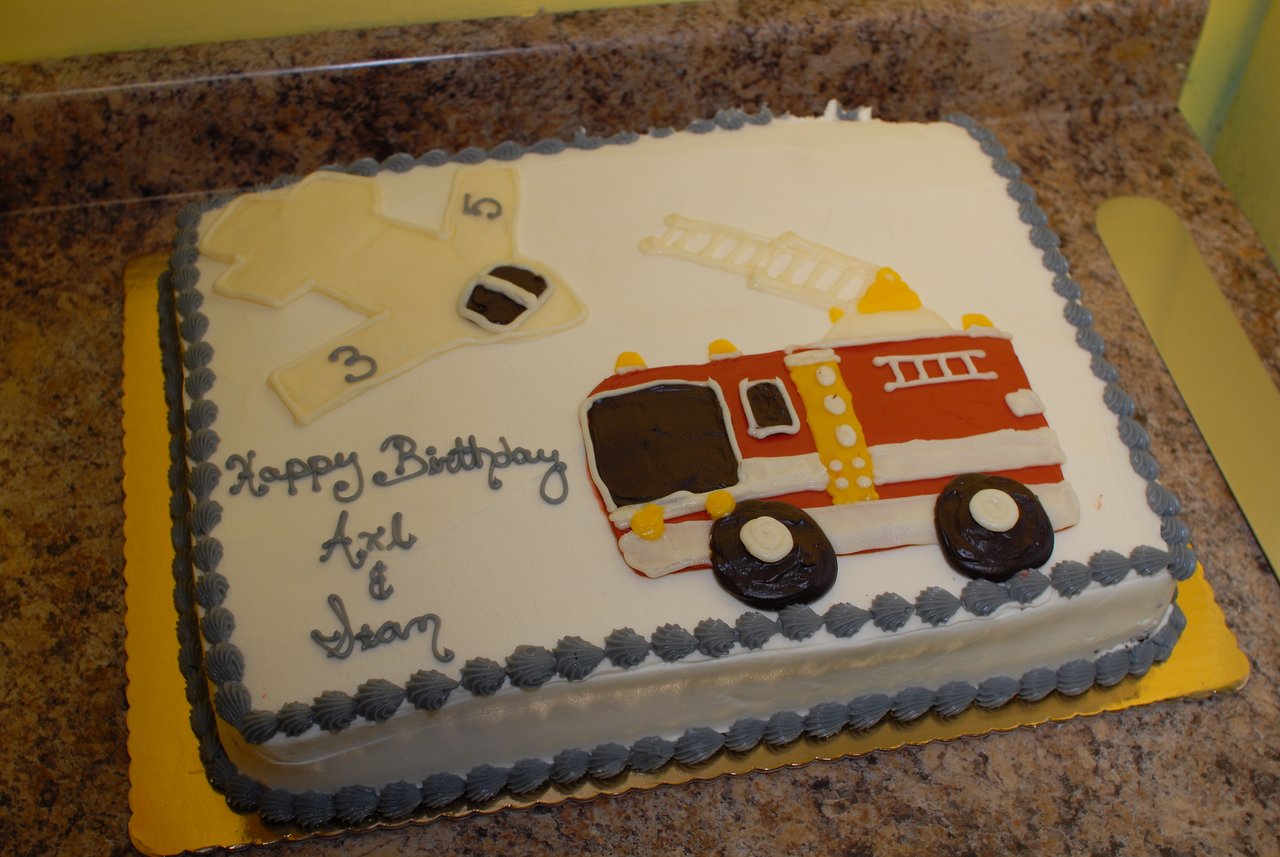 A rectangular birthday cake with a fire truck and firefighter-themed decorations, featuring "Happy Birthday Axl &amp; Sean" written in icing.