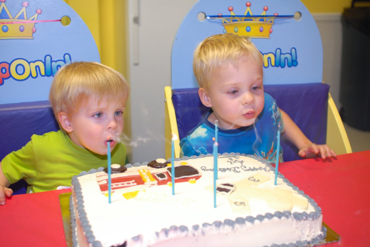 Two young children blow out candles on a decorated birthday cake while sitting at a table.