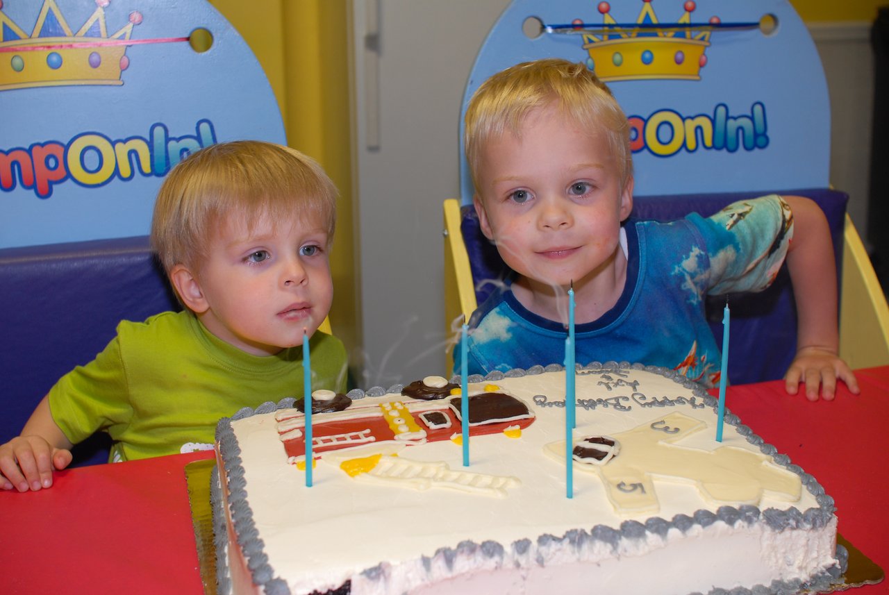 Two young children sit at a table with a birthday cake, blowing out blue candles.