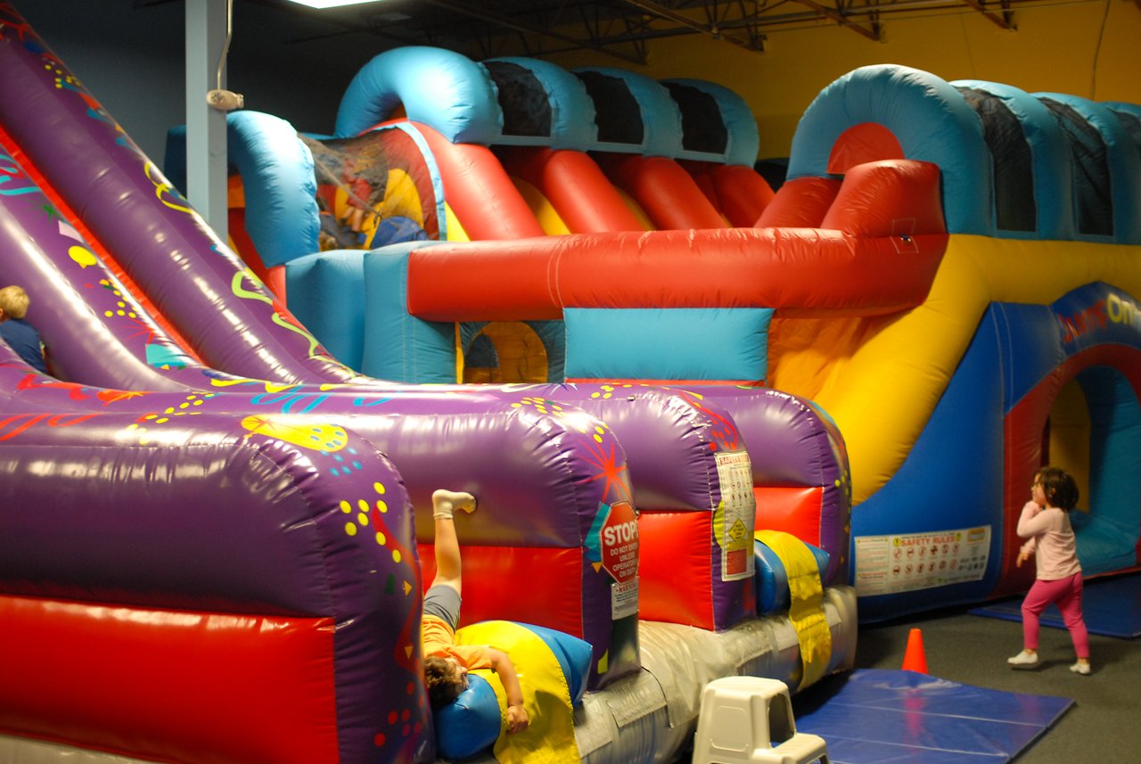 Children playing on a colorful inflatable bounce house with slides and obstacles at an indoor party venue.