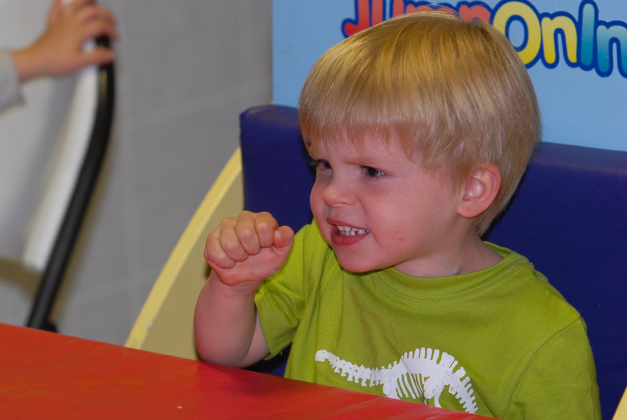 A young child in a green shirt makes a playful, determined expression while raising a fist at a table.
