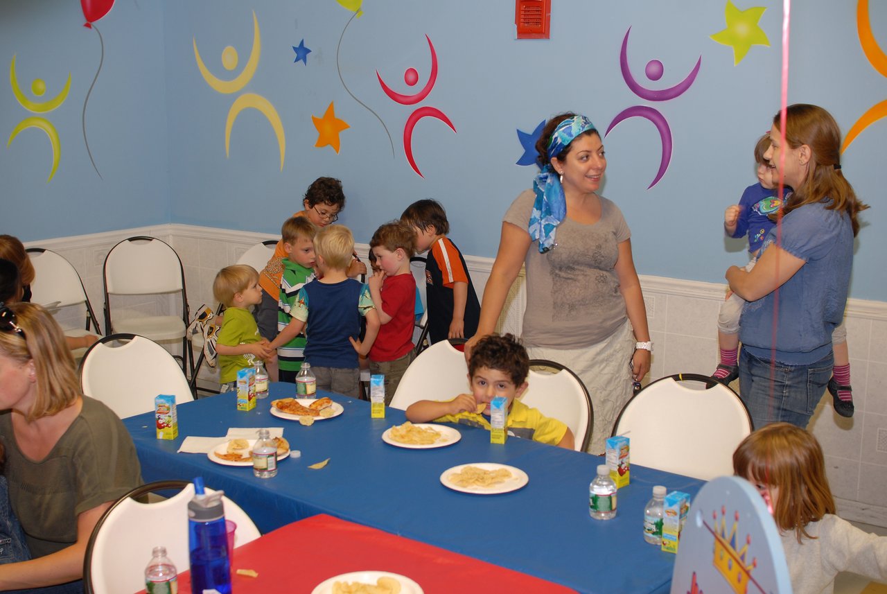 Children and adults gather at a birthday party, some chatting while others sit at a table with food and drinks.