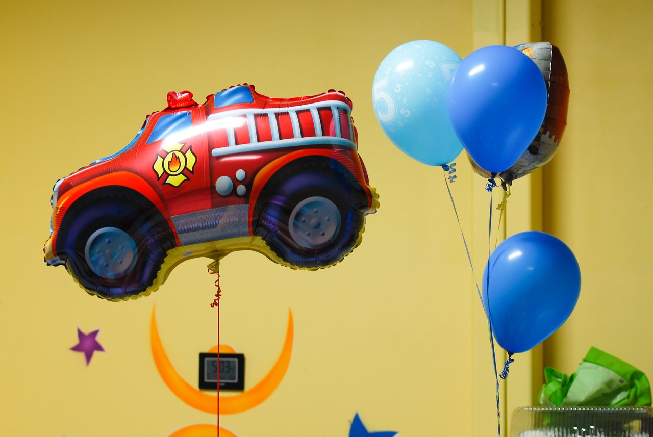 A red fire truck balloon and a cluster of blue balloons float against a yellow wall at a birthday party.