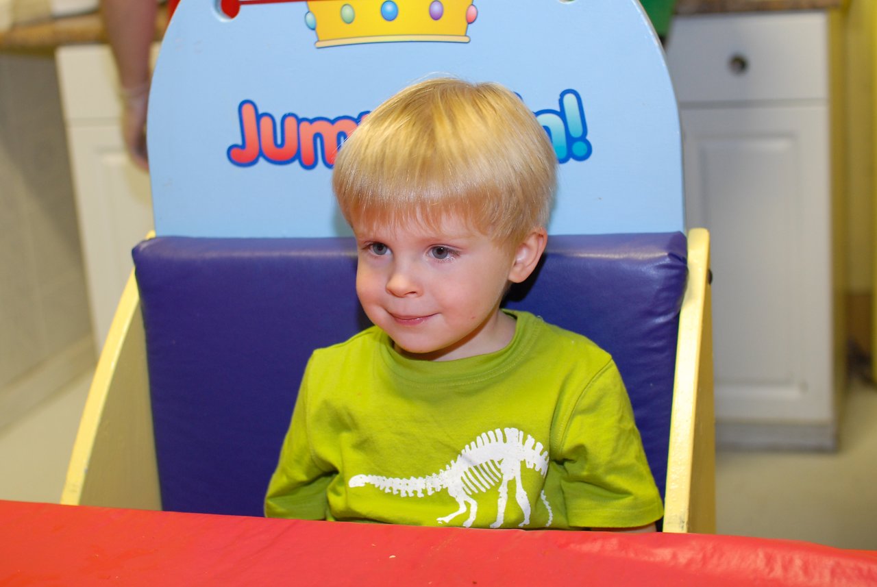 A young child in a green shirt sits at a table, waiting for birthday cake with a happy expression.