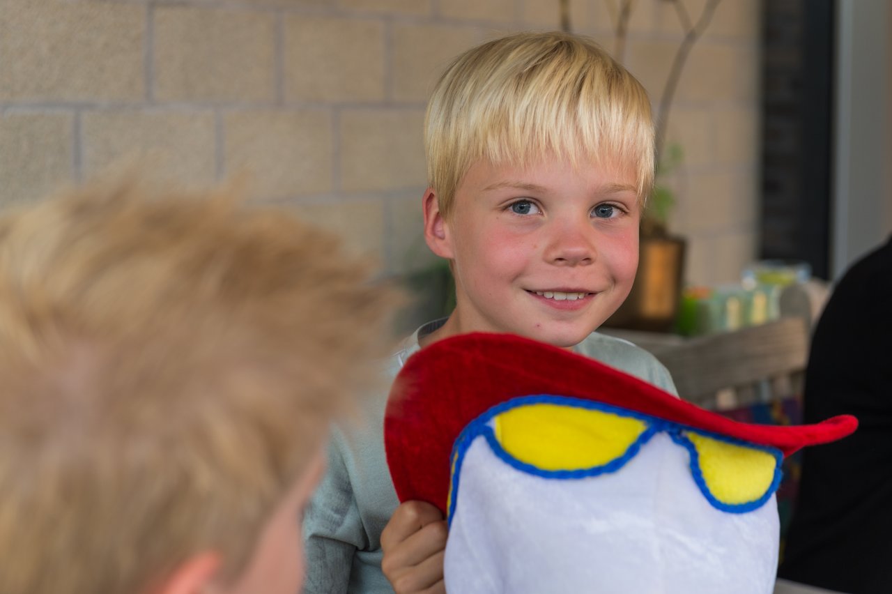 A smiling boy holds a colorful plush hat at his birthday celebration, with another child in the foreground.