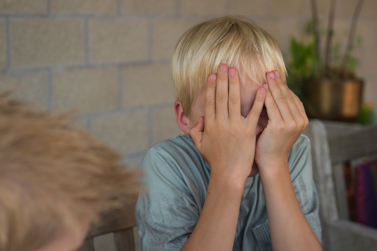 A young boy with blonde hair covers his face with both hands, possibly playing or reacting to something.