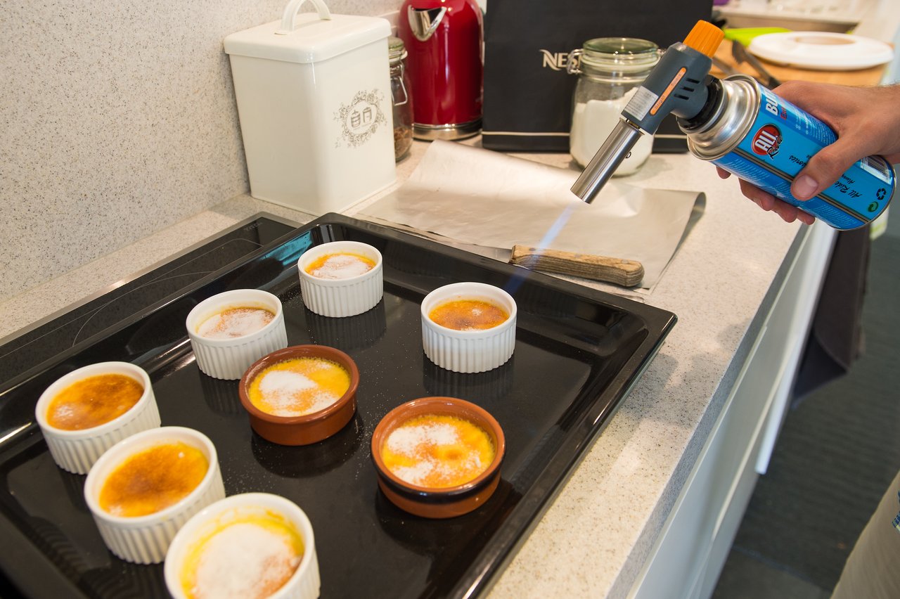 A person uses a kitchen torch to caramelize sugar on multiple crème brûlées arranged on a black tray.