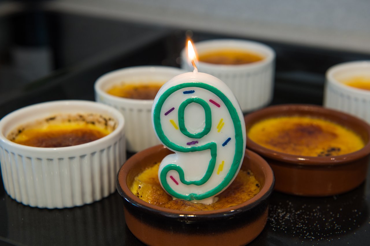 A lit number nine candle sits on crème brûlée, surrounded by more ramekins of the dessert for a birthday celebration.