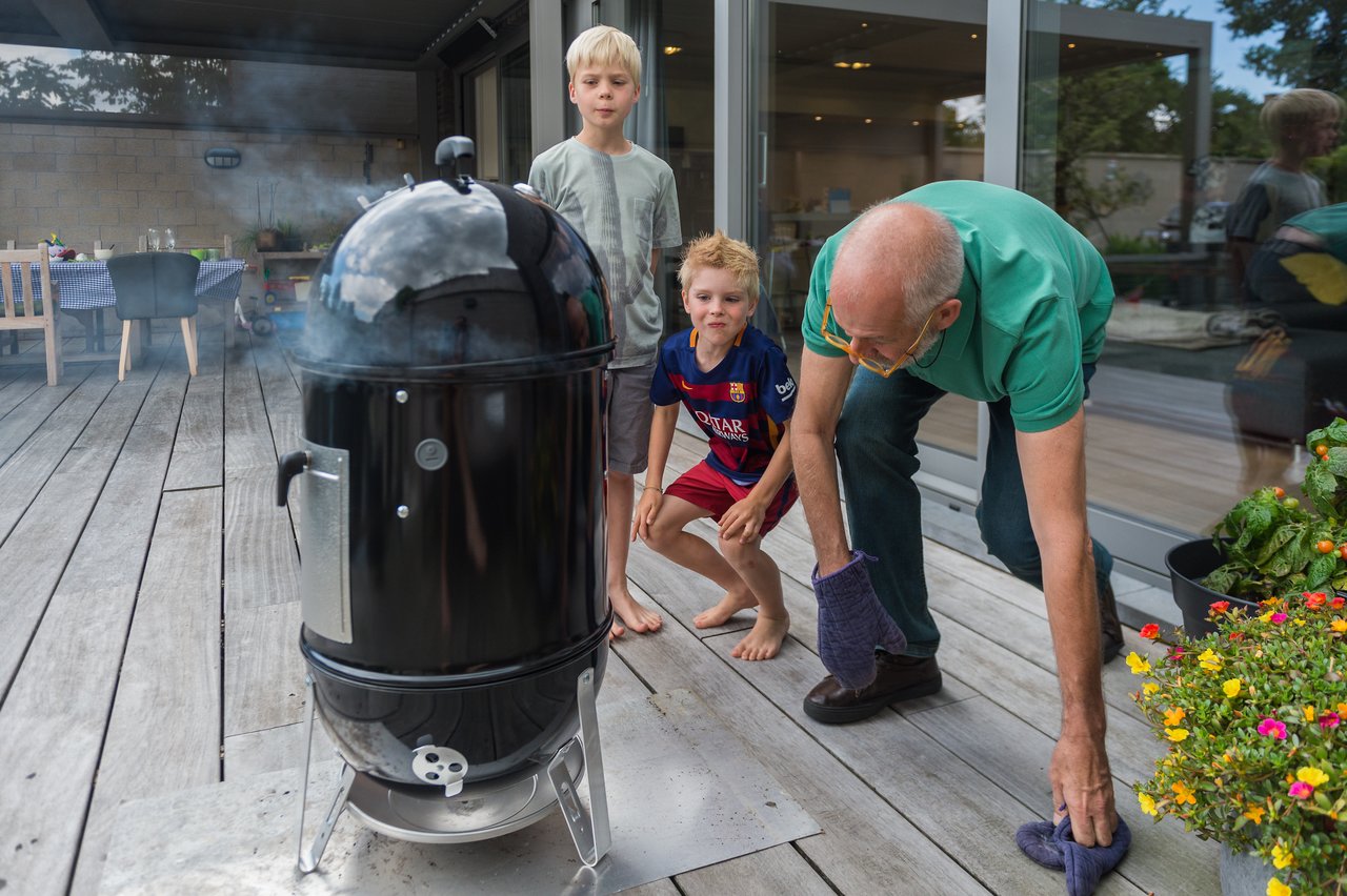 An older man and two children watch a smoking grill on a wooden deck, preparing food together.