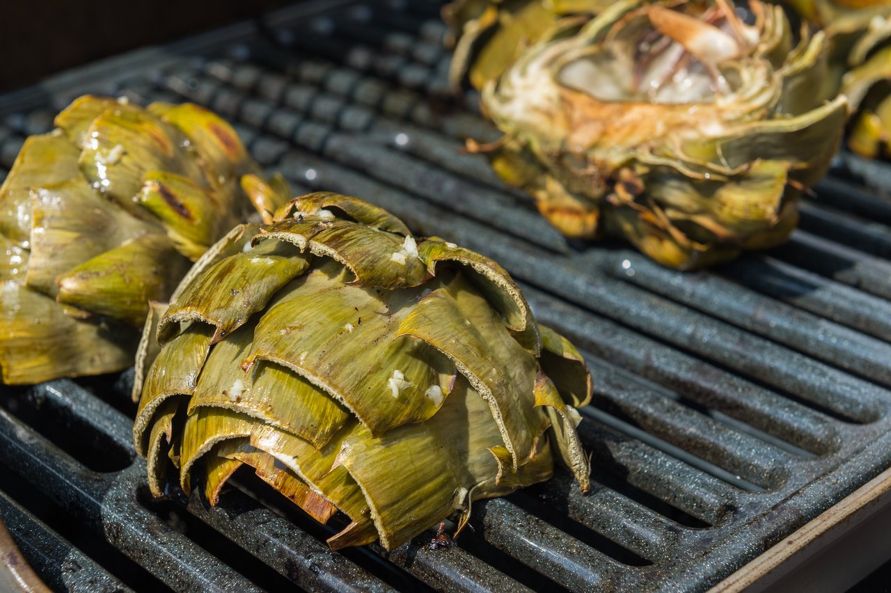 Artichokes cooking on a grill, with some cut open and seasoned.
