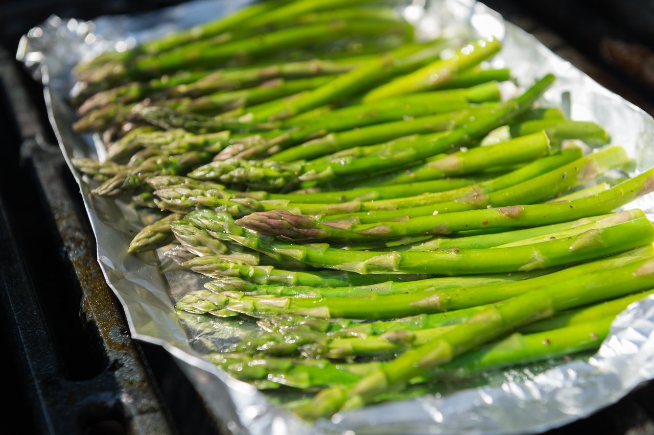 Fresh asparagus spears seasoned and placed on aluminum foil, grilling over an open flame.