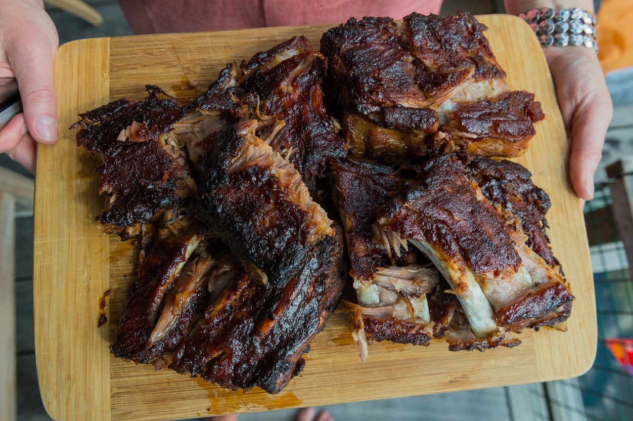 A person holds a wooden cutting board filled with freshly grilled and smoked ribs, ready to be served.