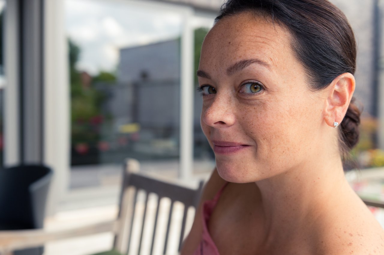 A woman with dark hair and earrings looks at the camera with a slight smile in an outdoor setting.