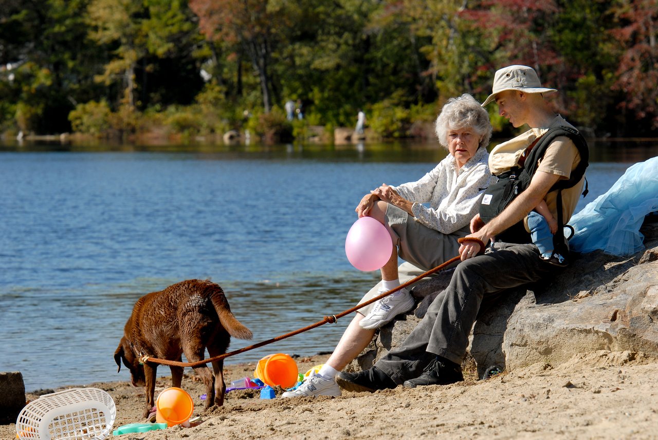 An elderly woman and a man with a baby sit by the water, holding a balloon and a dog leash.