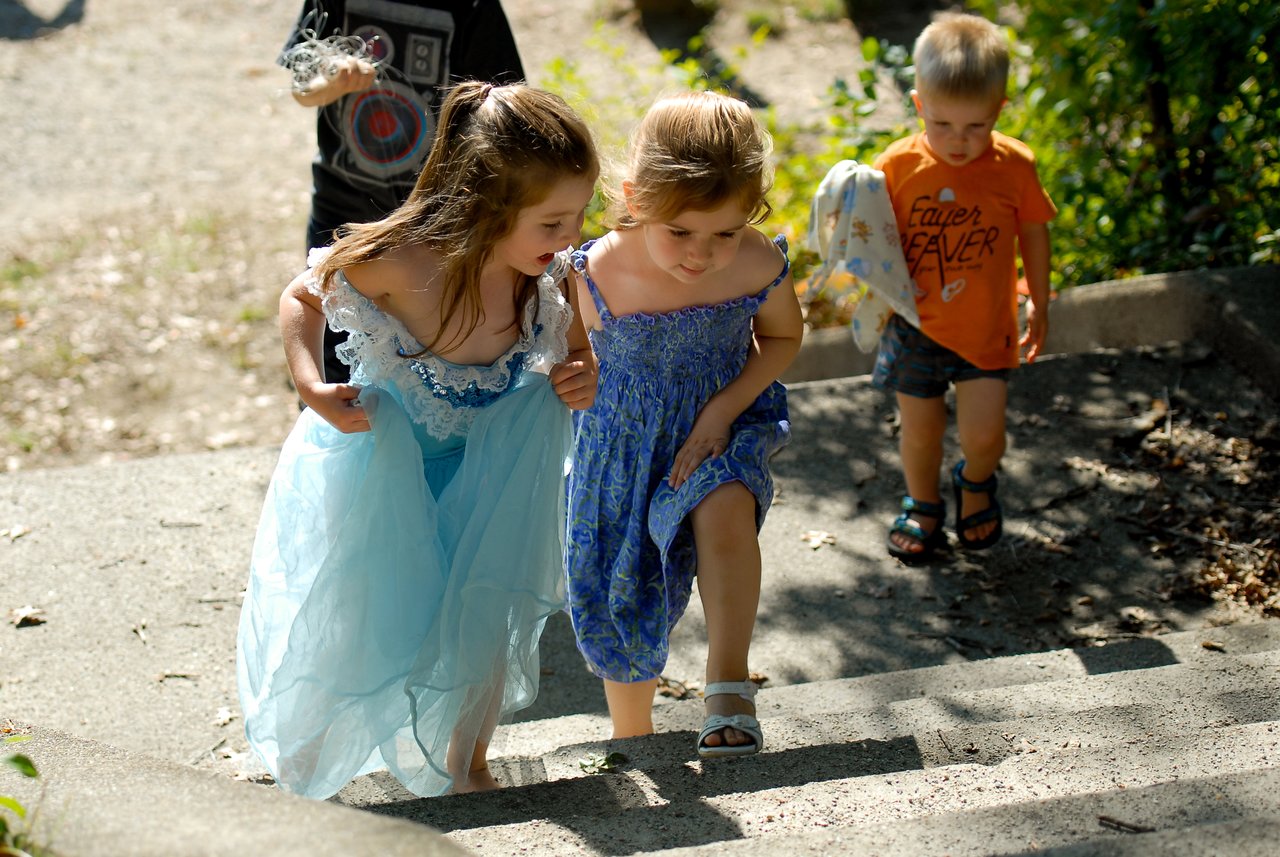 Two young girls in dresses walk up outdoor steps, followed by two children, one holding a stuffed toy.