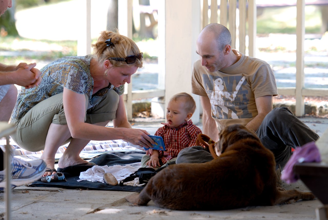A woman shows a small book to a baby while a man and a dog sit nearby.