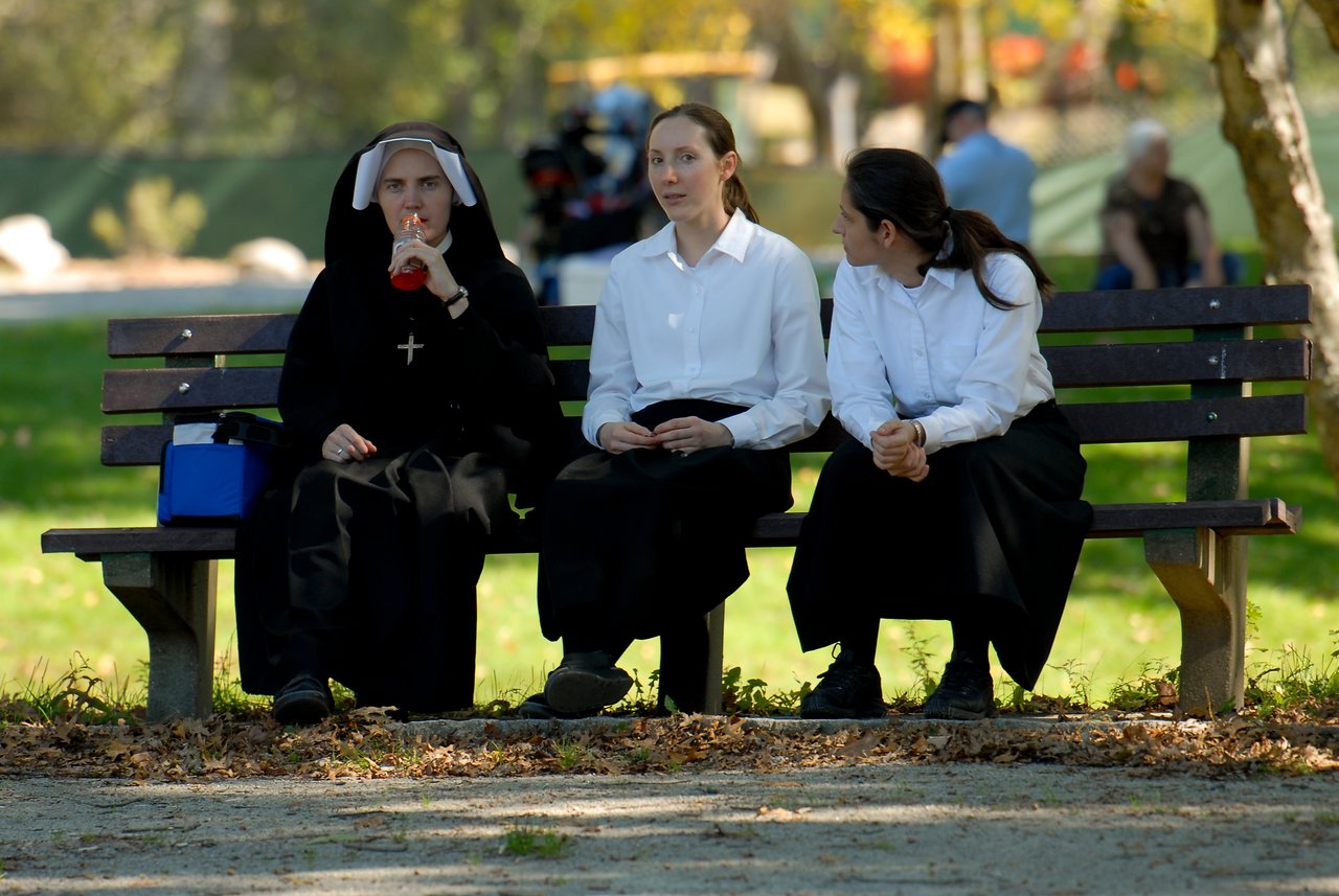 Three women in religious attire sit on a park bench, one drinking from a bottle while the others talk.