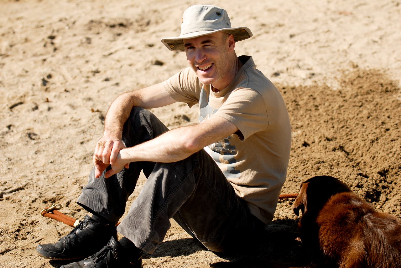 A man wearing a hat sits on sandy ground, smiling, with a brown dog lying nearby.