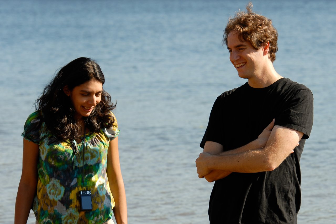Two people stand near the water, smiling and talking.