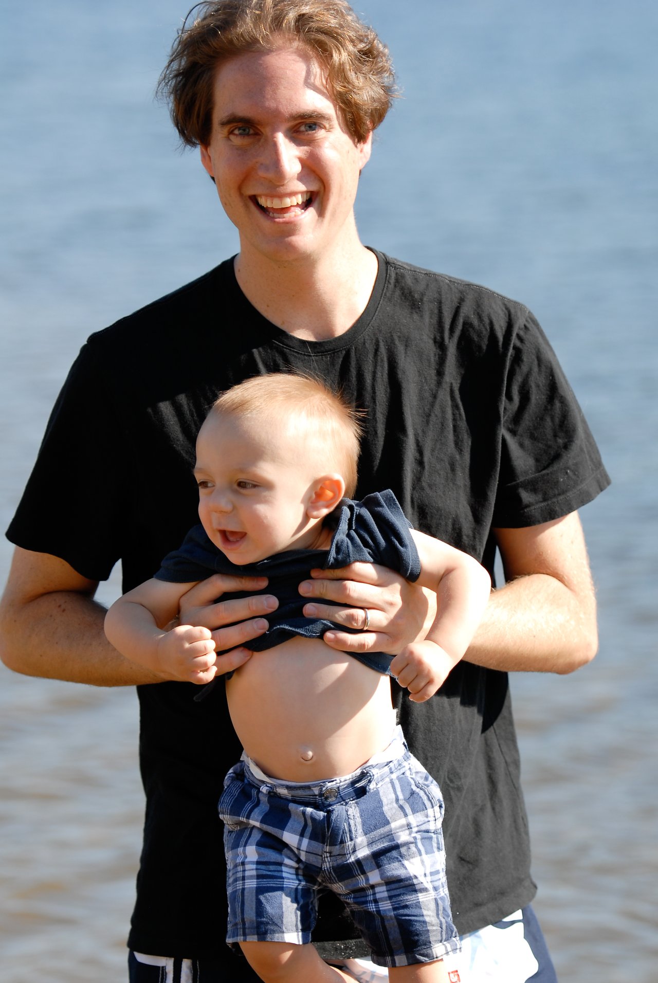 A man in a black shirt holds a smiling baby in plaid shorts near the water.