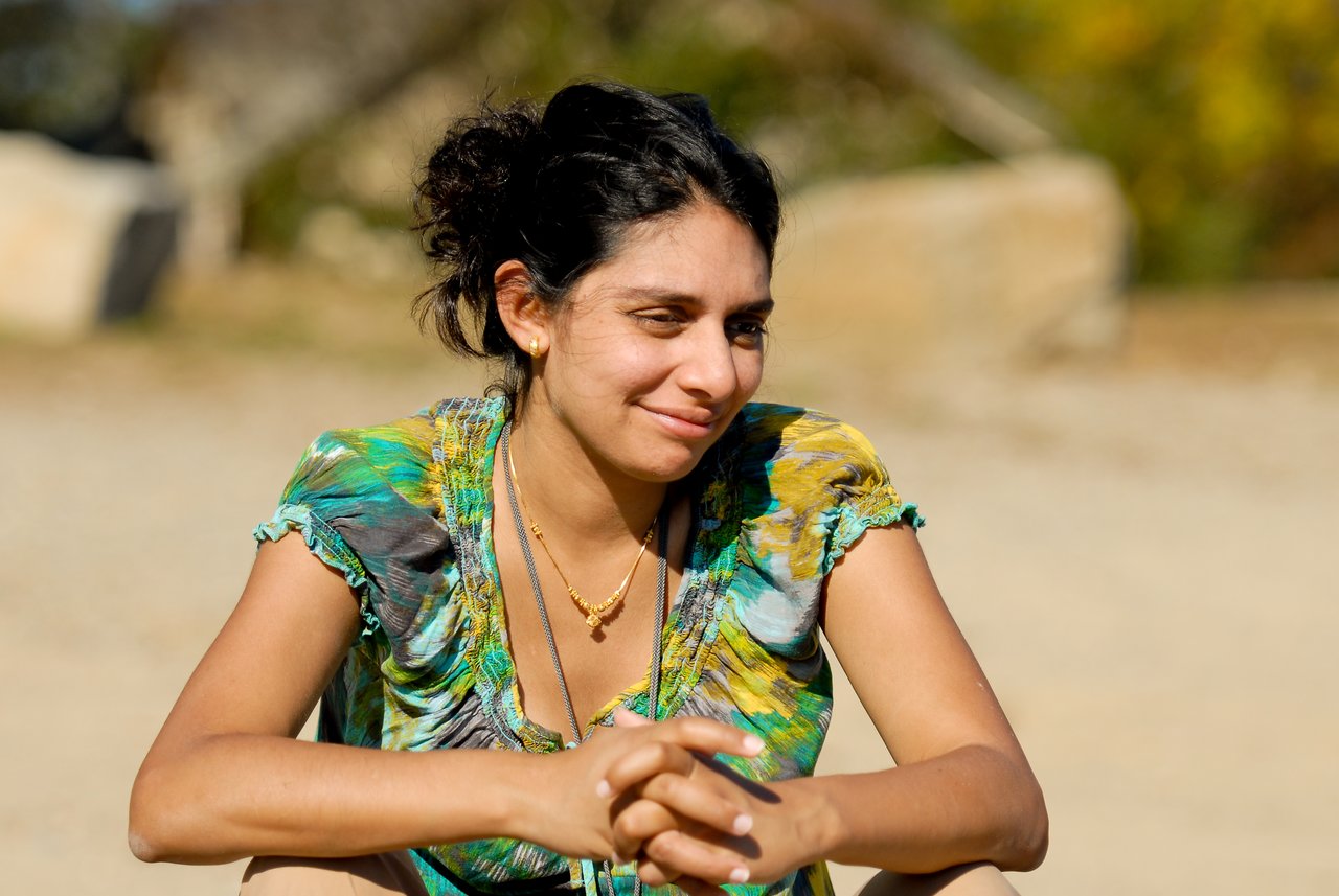A woman in a colorful top sits outdoors, smiling slightly and looking off to the side.