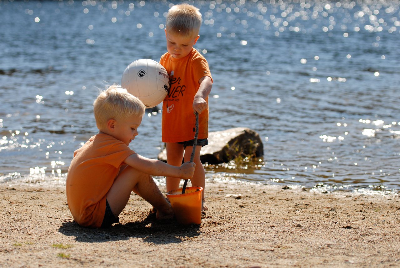 Two young children in orange shirts play with sand and a bucket near the water at a beach.
