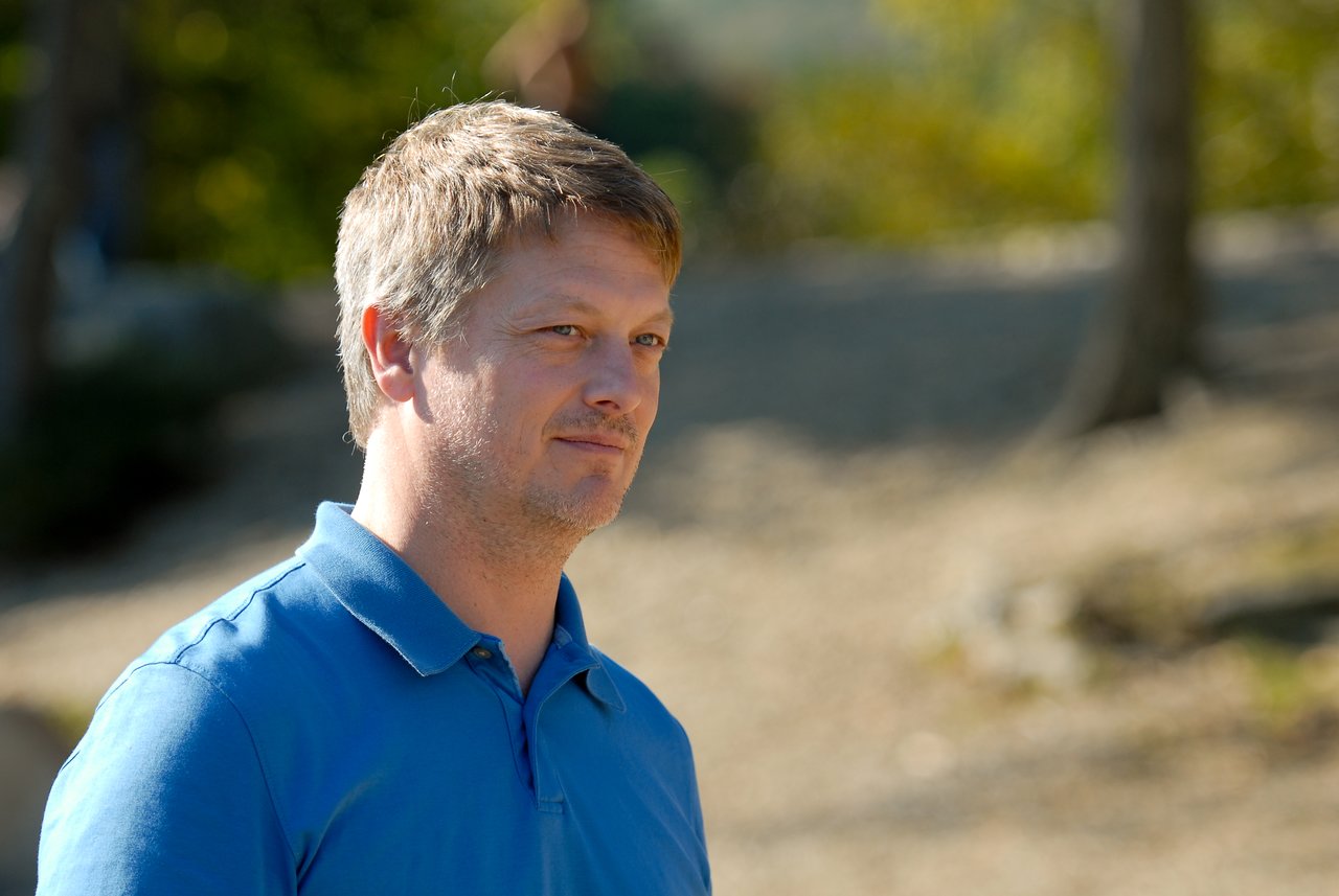 A man in a blue polo shirt stands outdoors, looking off to the side with a neutral expression.